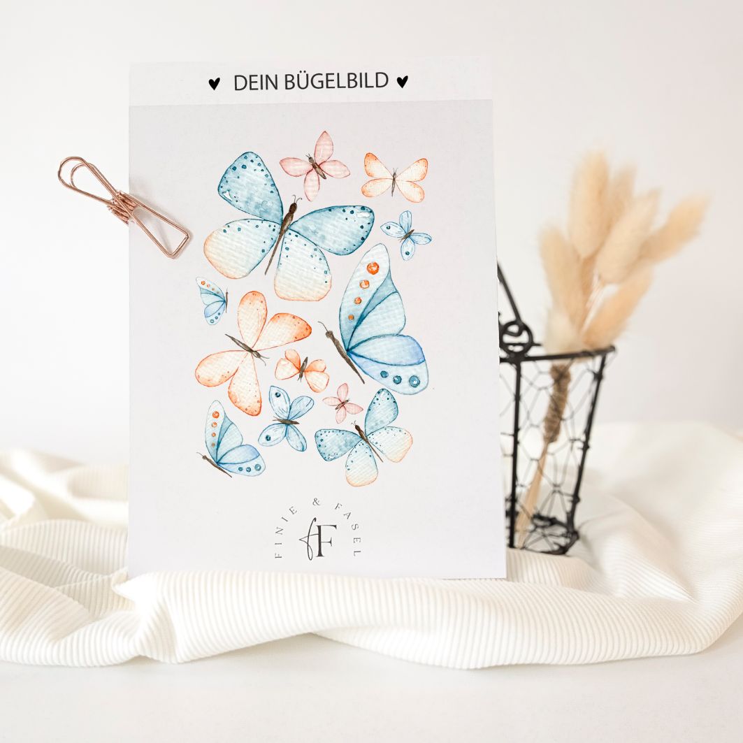 Bügelbild mit Schmetterlingsmotiv in Aquarell-Optik auf weißem Hintergrund, präsentiert auf einem Stoff mit Deko im Hintergrund – exklusiv bei Finie & Fasel
