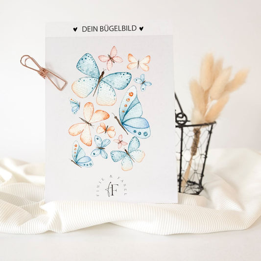 Bügelbild mit Schmetterlingsmotiv in Aquarell-Optik auf weißem Hintergrund, präsentiert auf einem Stoff mit Deko im Hintergrund – exklusiv bei Finie & Fasel