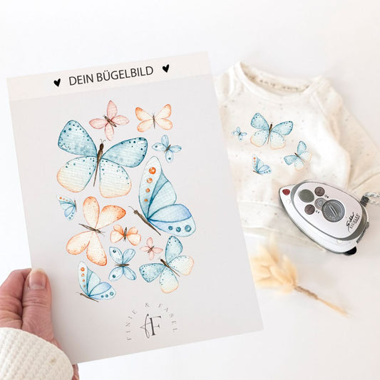 Bügelbild mit handgemalten Schmetterlingen in sanften Farben, daneben ein Pullover mit aufgebügeltem Motiv – ideal zum Personalisieren von Kinderkleidung.